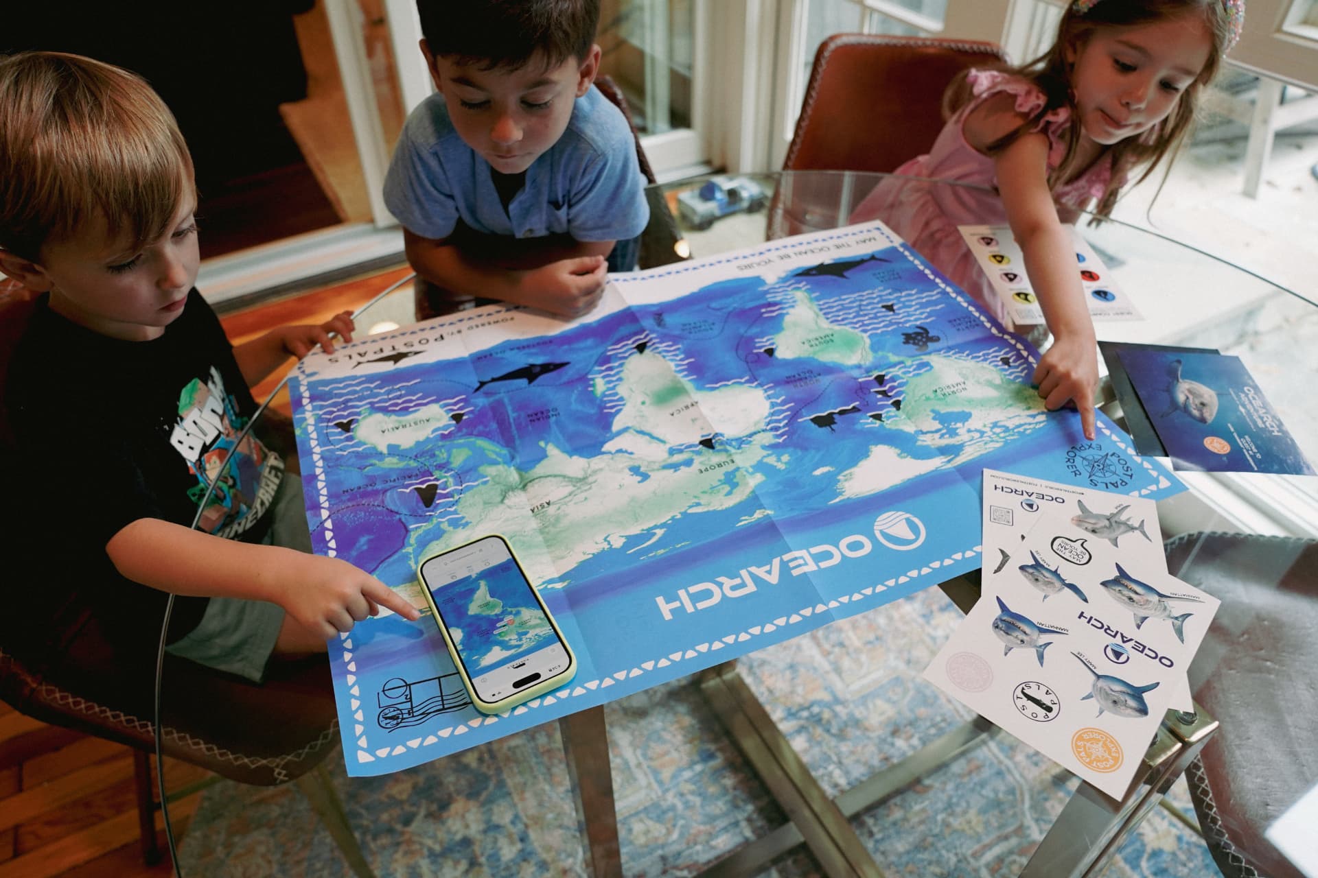 Three children leaning over a large POSTPALS OCEARCH shark tracking map, placing stickers while one holds a phone showing a Quest